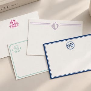 May include: Four rectangular white stationery cards featuring personalized monograms. One card has a pink ornate monogram, one has a purple diamond monogram with lines, one has a teal monogram with a border, and one has a navy blue circular monogram.