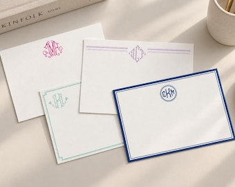 May include: Four rectangular white stationery cards featuring personalized monograms. One card has a pink ornate monogram, one has a purple diamond monogram with lines, one has a teal monogram with a border, and one has a navy blue circular monogram.