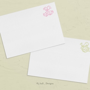May include: Two blank white note cards with a decorative floral monogram in pink and green. The monogram is the letter "S" with a flourish.