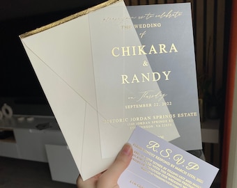 May include: A white envelope with a gold border and a clear plastic invitation card with gold lettering. The invitation card reads "Please join us to celebrate the wedding of Chikara & Randy on Thursday, September 22, 2022 at Historic Jordan Springs Estate, 1150 Jordan Springs Rd,  Steffenson, VA 22656". A white RSVP card with gold lettering is also included.