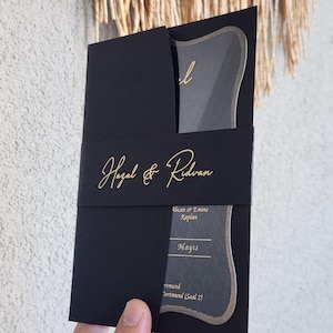 Könnte beinhalten: Schwarze Hochzeitseinladung mit goldener Schrift. Die Einladung zeigt die Namen "Hazal & Ridvan" auf einem schwarzen Band. Sie enthält einen transparenten Einsatz mit goldener Umrandung und zusätzlichem Text.
