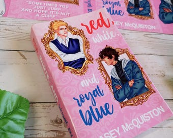 Può includere: Un libro rosa intitolato "Red, White, and Royal Blue" di Casey McQuiston. La copertina presenta illustrazioni di due uomini in cornici dorate ornate. Il titolo è scritto in corsivo rosso e blu, con sfondo bianco.