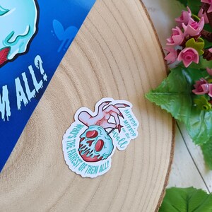 May include: A sticker with a cartoon skull and a red apple core, a hand with long red nails, and the text "Mirror, mirror on the wall, who's the fairest of them all?" The sticker is on a wooden surface.