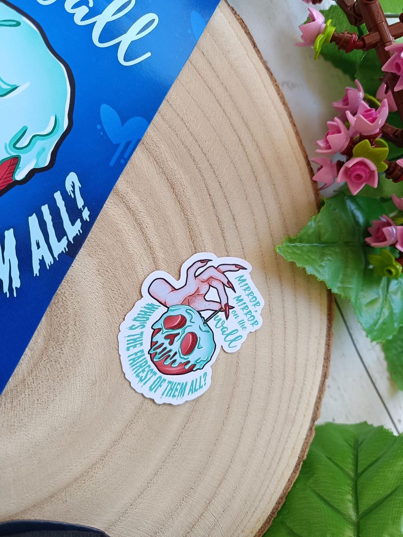 May include: A sticker with a cartoon skull and a red apple core, a hand with long red nails, and the text "Mirror, mirror on the wall, who's the fairest of them all?" The sticker is on a wooden surface.