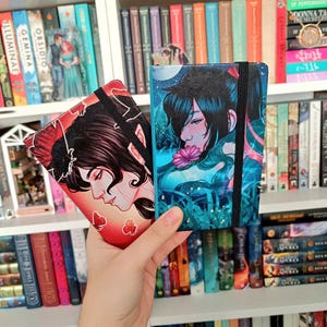 Puede incluir: Dos pequeños diarios de tapa dura con cubiertas coloridas que presentan ilustraciones de personas con cabello largo y oscuro. El diario de la izquierda tiene una cubierta roja con un diseño de mariposa y el diario de la derecha tiene una cubierta azul con un diseño de flor.