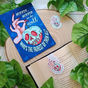 May include: A blue bookmark and sticker featuring a poison apple design. The bookmark reads "Mirror, Mirror on the wall, Who's the fairest of them all?" An open book with the bookmark inside. Green leaves surround the items.