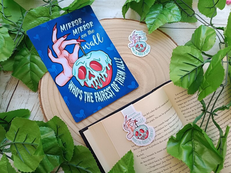 May include: A blue bookmark and sticker featuring a poison apple design. The bookmark reads "Mirror, Mirror on the wall, Who's the fairest of them all?" An open book with the bookmark inside. Green leaves surround the items.