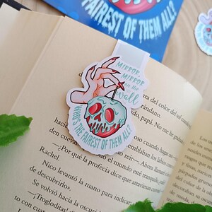May include: A bookmark with a hand holding a poison apple design, featuring the text "Mirror, Mirror on the wall" and "Who's the fairest of them all?" It's placed in a book. A matching sticker is also visible.