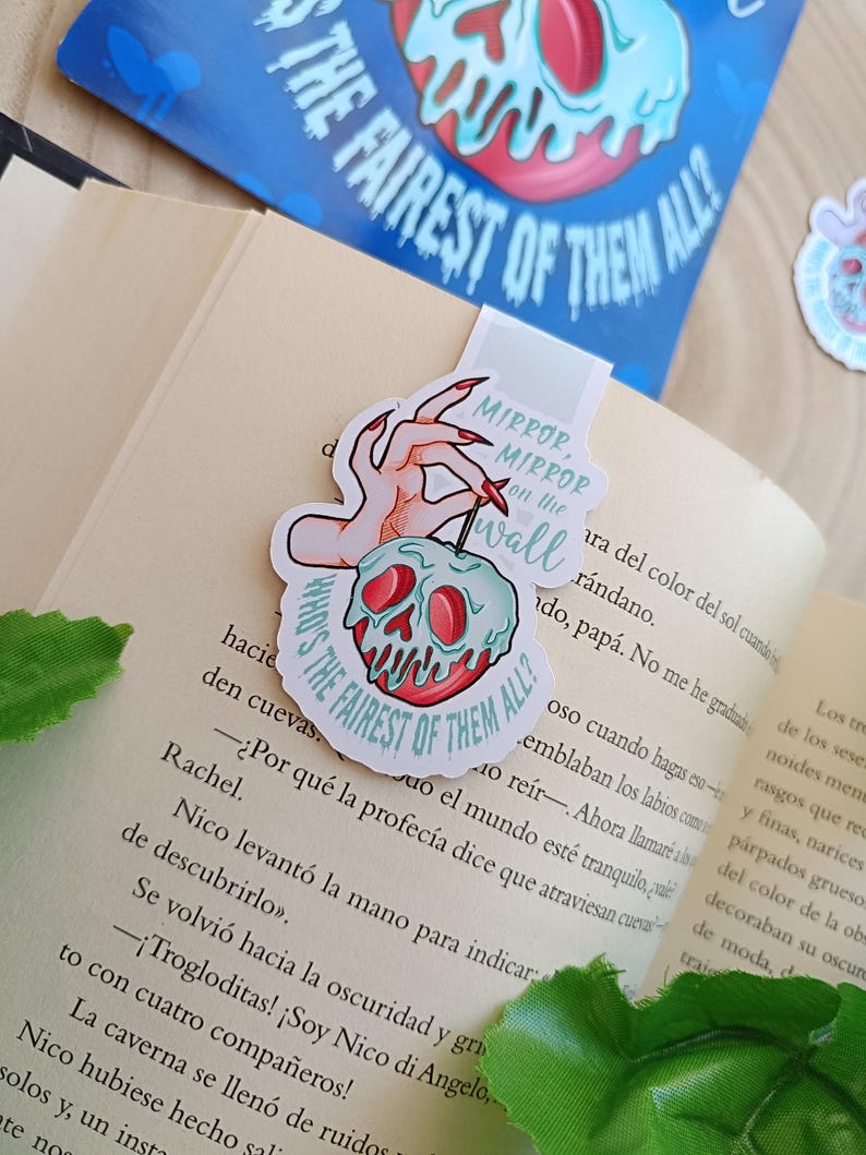 May include: A bookmark with a hand holding a poison apple design, featuring the text "Mirror, Mirror on the wall" and "Who's the fairest of them all?" It's placed in a book. A matching sticker is also visible.