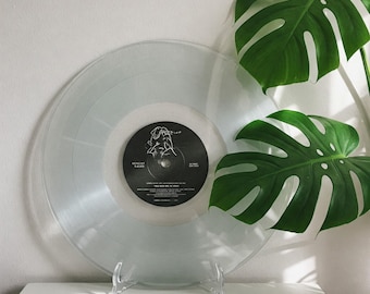 Könnte beinhalten: Eine transparente Vinyl-Schallplatte mit einem schwarzen Label, auf dem eine weiße Linienzeichnung einer Person zu sehen ist. Die Schallplatte steht auf einem transparenten Acryl-Ständer. Im Hintergrund befindet sich eine grüne Monstera-Pflanze.