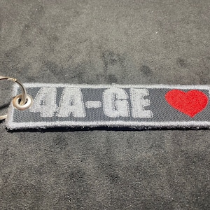 May include: A black key chain with a silver metal ring. The keychain has a white embroidered text that says "4A-GE" and a red heart.