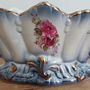 Pode incluir: Uma tigela oval de cerâmica com borda recortada e acabamento dourado. O exterior apresenta um degradê azul e branco com detalhes florais em relevo e acentos dourados. Um motivo central exibe rosas cor-de-rosa e folhas verdes na frente.