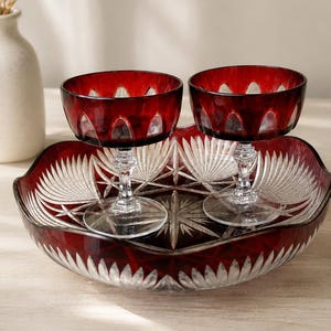 Può includere: Un set di due coppe in cristallo rosso intenso con steli e basi trasparenti, poggiate all'interno di una grande ciotola in cristallo rosso coordinata. Il set presenta intricati motivi intagliati, tra cui disegni a stella e bordi smerlati.