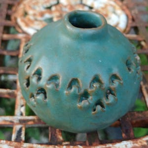 Può includere: Un piccolo vaso in ceramica color verde acqua, di forma rotonda con collo e apertura stretti. La superficie presenta un motivo ripetitivo di piccole rientranze triangolari che rivelano una tonalità marrone scuro nell'argilla testurizzata.