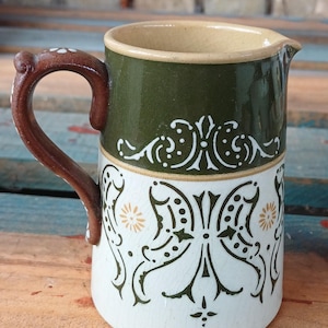Può includere: Una brocca in ceramica con una fascia superiore verde scuro e un corpo inferiore bianco decorato con complessi motivi floreali in verde scuro e beige. L'oggetto presenta un manico marrone con accenti bianchi e una finitura smaltata craquelé.