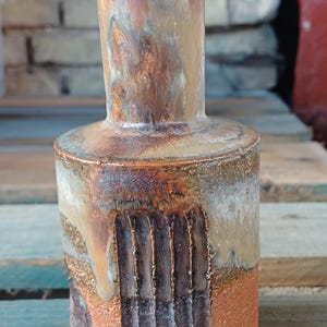 Op de afbeelding: Een cilindrische keramische vaas met een smalle hals, voorzien van een getextureerd oppervlak met verticale groeven. De afwerking toont een mix van aardse bruine, roestkleurige en gedempte grijstinten met een druipend glazuureffect bovenaan.