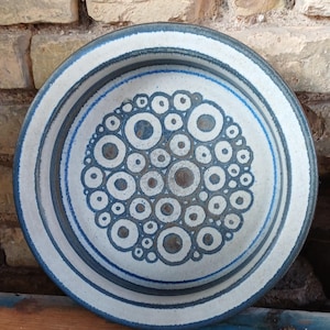 Peut inclure: Un bol en céramique rond présentant un émail blanc cassé moucheté avec un motif central de cercles concentriques bleu foncé et marron. Le bord est accentué par une bande bleu foncé et la base intérieure est remplie de cercles de tailles variées.