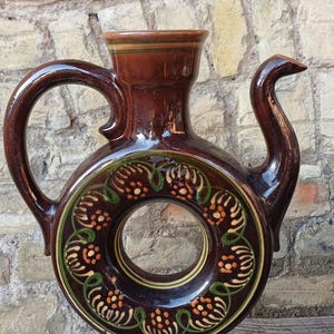 Könnte beinhalten: Ein dunkelbraun glasierter Keramikkrug mit einem kreisförmigen, ringförmigen Körper und einem zentralen Loch. Das Gefäß ist mit handgemalten grünen und orangefarbenen Blumenmotiven verziert und verfügt über einen geschwungenen Henkel, einen schmalen Ausguss und einen ausgestellten Fuß.
