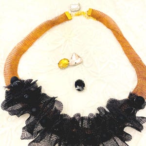 May include: A statement necklace featuring a gold-toned mesh chain with a black lace and bead embellishment. The necklace is adorned with a black gem and three additional decorative gems in yellow and clear colors.