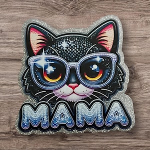 May include: A black cat wearing sunglasses with the word "MAMA" in a blue, glittery font. The cat has a pink nose and ears, and the image has a silver glitter border. The cat's glasses have a sparkling effect.
