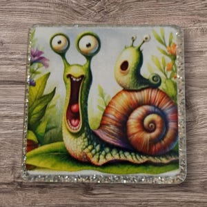 May include: Square coaster featuring a whimsical illustration of two snails. The larger snail has a wide open mouth and large eyes. The smaller snail sits on top of the larger snail's shell. The background includes green leaves and flowers.
