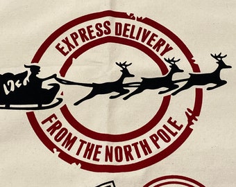 May include: Cream-colored fabric with a red and black graphic of Santa Claus in his sleigh, pulled by reindeer. The text reads "EXPRESS DELIVERY FROM THE NORTH POLE" in a circular stamp design. Additional text is visible.