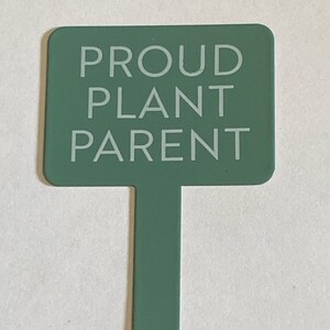 Könnte beinhalten: Ein grüner Pflanzenmarker mit dem Text "PROUD PLANT PARENT" in weißen Buchstaben.