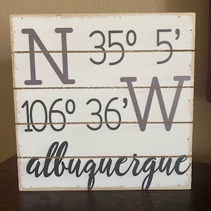 Puede incluir: Un letrero de madera blanco con letras negras. El letrero dice "N 35° 5' 106° 36' W albuquerque".