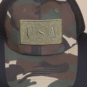 May include: Camouflage baseball cap with a dark mesh back. The front of the cap features a green rectangular patch with the letters "USA" embroidered in a matching green thread. The cap has a curved brim and a classic six-panel design.