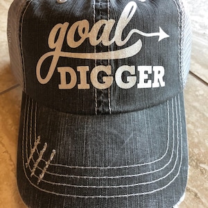 May include: A charcoal gray trucker hat with the words "goal DIGGER" in white script and block letters. The hat has a distressed look with a mesh back and a curved brim with white stitching.