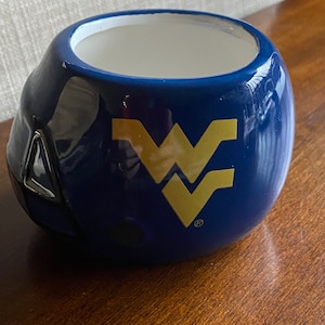 May include: Blue ceramic mug shaped like a football helmet with a gold West Virginia University logo. The mug has a white interior and a black rim.