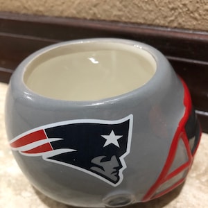 Pode incluir: Uma caneca de cerâmica cinza em forma de capacete de futebol americano com o logotipo dos New England Patriots. O logotipo é uma estrela vermelha, branca e azul com uma asa azul e um contorno branco.