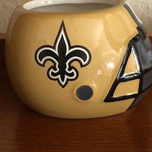 May include: A ceramic football helmet planter in gold with a black fleur-de-lis logo. The helmet has a black and white stripe design on the side.