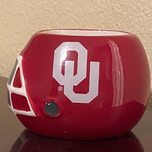May include: A red and white ceramic Oklahoma Sooners football helmet mug. The mug has the Oklahoma Sooners logo on the front and a registered trademark symbol.