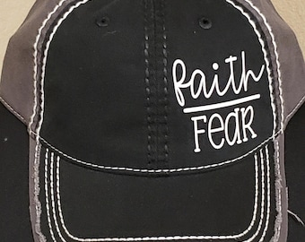 Puede incluir: Gorra de béisbol negra con visera desgastada y paneles grises. La gorra presenta costuras blancas y las palabras "faith" sobre "fear" en escritura blanca.