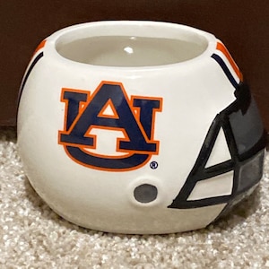 May include: White ceramic football helmet mug with an orange and blue "AU" logo. The mug has a black facemask and a white interior.