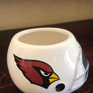 May include: White ceramic planter shaped like a football helmet with a red cardinal logo. The planter has a black rim and a black football.
