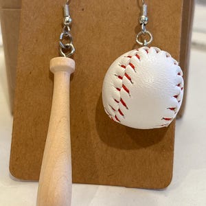 Op de afbeelding: Baseball-oorbellen. Eén oorbel heeft een miniatuur houten honkbalknuppel, de andere een witte honkbal met rode stiksels. Beide hebben zilverkleurige oorhaakjes.