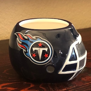 Pode incluir: Uma caneca de cerâmica azul e branca em forma de capacete de futebol americano com o logotipo dos Tennessee Titans. O logotipo apresenta uma chama azul com um "T" branco dentro de um círculo com duas estrelas brancas.