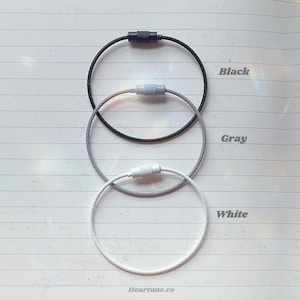 May include: Three keychains in black, gray, and white, each with a metal cable loop and a screw-on clasp. The colors are labeled on the right side of the image. The keychains are arranged vertically on a lined paper background.