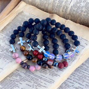 May include: Several beaded bracelets with a variety of stone beads, including black lava rock, tiger's eye, and pink stones. Each bracelet features a silver cross charm. The bracelets are displayed on a vintage book.