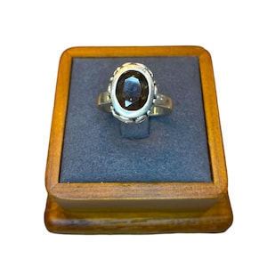 May include: A silver ring with a brown oval gemstone set in a decorative bezel. The ring is displayed in a wooden ring box.