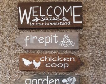 May include: Six wooden signs with white lettering and illustrations. The signs read: Welcome to our homestead, firepit, chicken coop, garden, outhouse, and beehives.