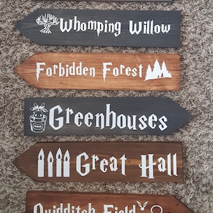 Puede incluir: Seis señales de madera en forma de flecha con texto blanco y gráficos. Las señales dicen: La cabaña de Hagrid, El sauce boxeador, El bosque prohibido, Los invernaderos, El Gran Comedor y El campo de Quidditch.