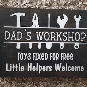 May include: A dark brown wooden sign with white text that reads "Dad's Workshop" with a row of tools above the text and another row of tools below the text. The sign also says "Toys Fixed For Free" and "Little Helpers Welcome".