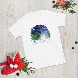 Puede incluir: Camiseta blanca con un coche azul volando a través de una escena de bosque nevado. El texto "Merry Christmas" está impreso debajo de la imagen.