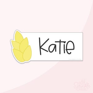 Könnte beinhalten: Ein rechteckiges weißes Namensschild mit dem Namen Katie in schwarzer, handschriftlicher Schrift. Auf der linken Seite des Schildes befindet sich eine Anordnung von fünf blassgelben, blütenblattförmigen Elementen in einem vertikalen Stapel.