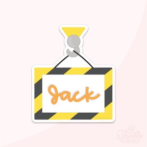 May include: A yellow and black construction sign with the name "Jack" written in orange. The sign is hanging from a grey hook.