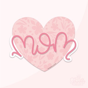 May include: A pink heart-shaped sticker with a floral pattern and the word "MOM" in a cursive font. The sticker has a white border and the text "THE SPRINKLE FACTORY" at the bottom. The background is a light pink.
