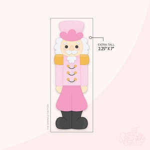 Puede incluir: Un cortador de galletas de cascanueces rosa y dorado. El cortador es extra alto, mide 5,7 cm por 17,8 cm. El cascanueces tiene un sombrero, uniforme y pantalón rosa, con charreteras doradas y botas negras. El texto "THE SPRINKLE FACTORY" está impreso en el lateral.